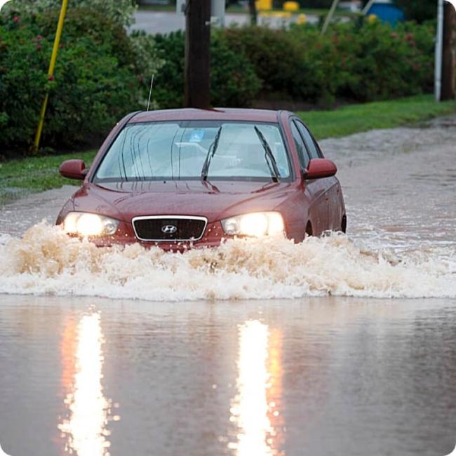 A red car drives through deep floodwaters, creating large splashes.