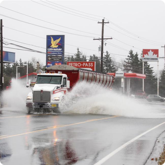 A large white tanker truck splashes through standing water on a road near gas stations.