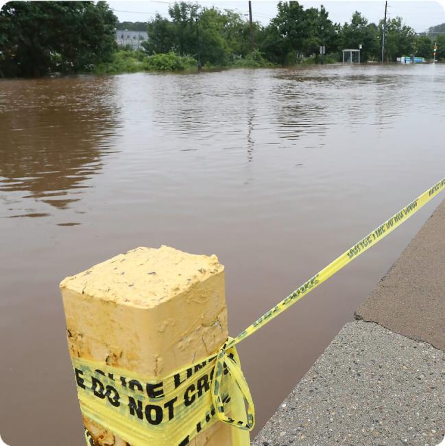 A "Police Line Do Not Cross" tape blocks a flooded street next to a yellow concrete post.