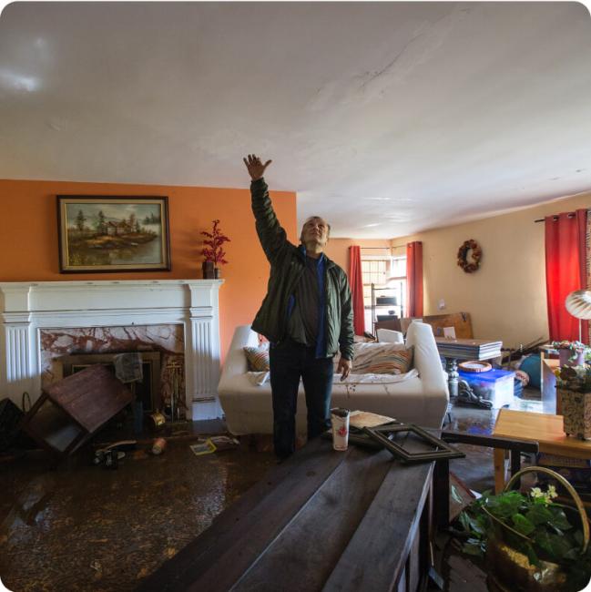 Man with hand raised points to a damaged ceiling in a flooded room.