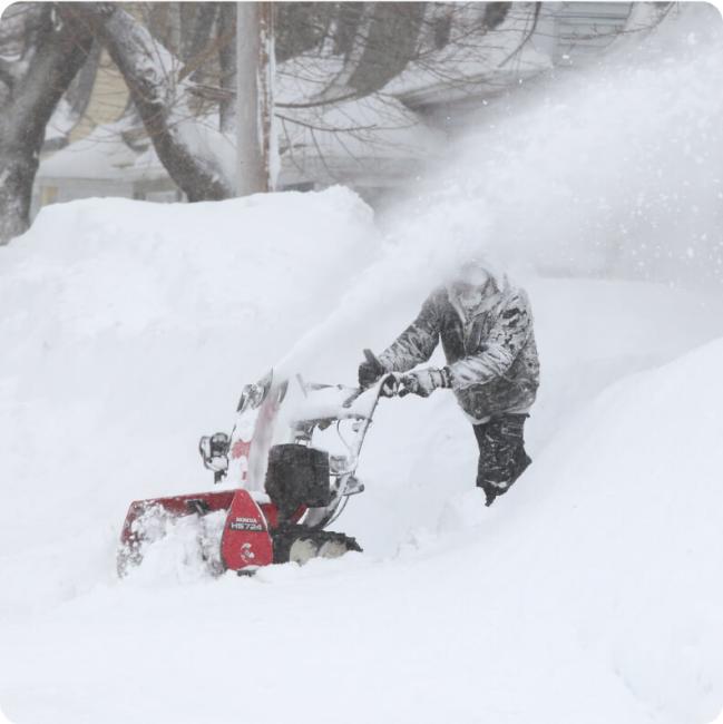 A person operates a red snow blower on a snow-covered residential street during a blizzard.