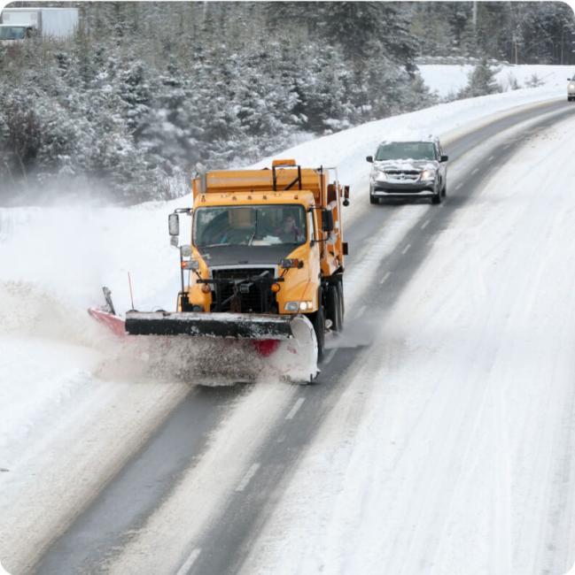 A yellow snow plow truck clears a snowy road, kicking up snow to the side.