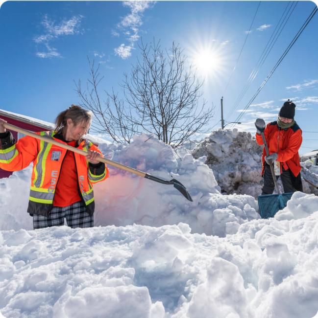 Two people shovel deep snow outside a house on a sunny winter day.